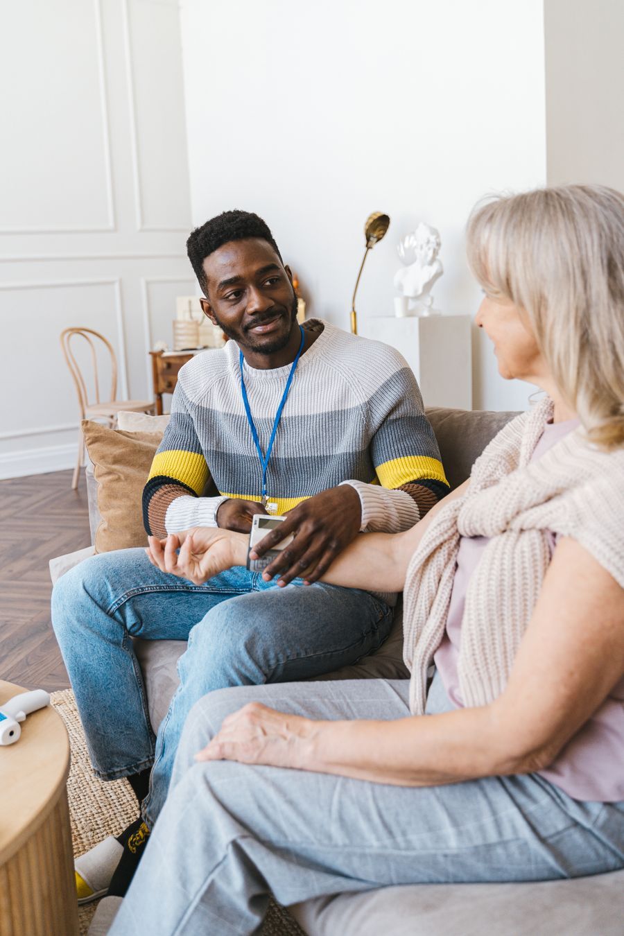 A health worker with a lanyard talking with an older woman on her sofa at home