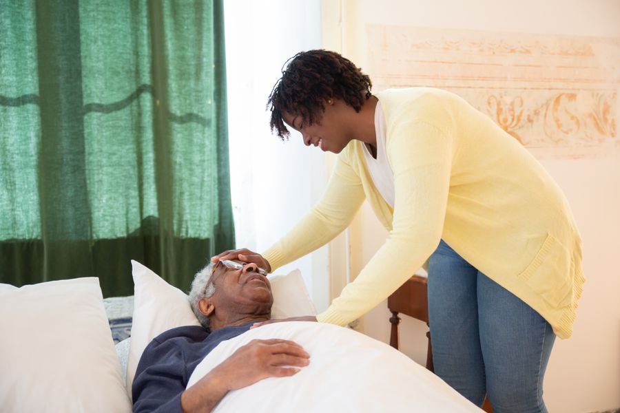 A carer gently checking the temperature of an elderly man resting in bed